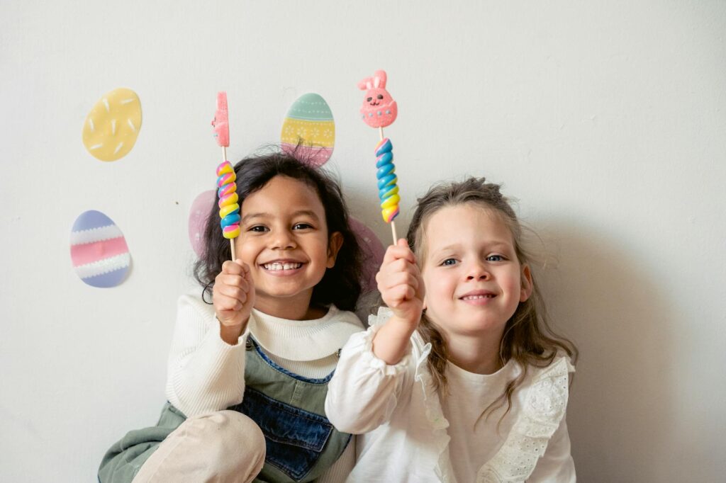 Cheerful multiracial children with sweet Easter bunny candies looking at camera while sitting on white background with egg decorations during holiday celebration