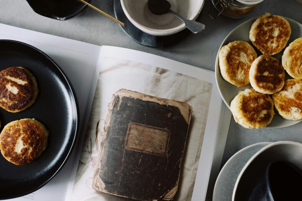 From above view of fresh hot buns served on plates near opened book on table with various kitchenware in light kitchen