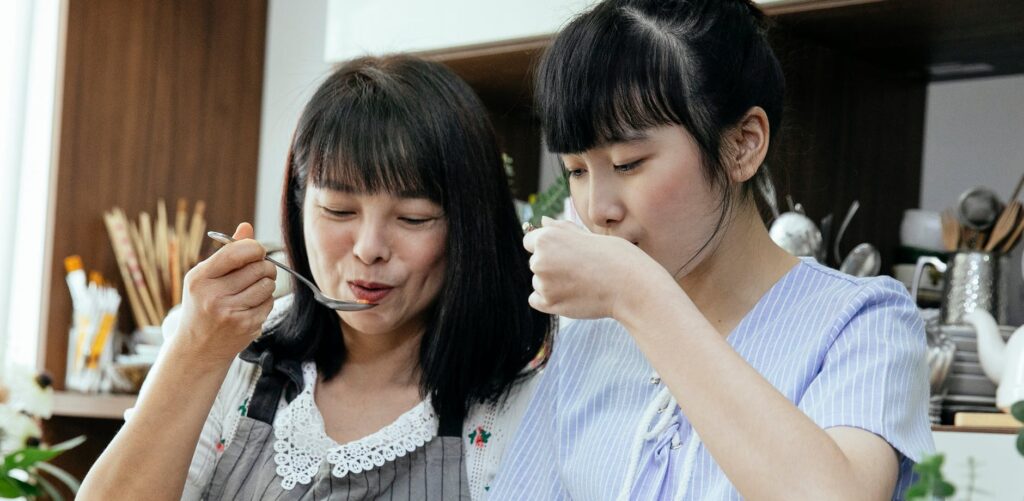 Asian mother and daughter with spoons tasting fresh meal and standing in kitchen with various cutlery while cooking lunch together