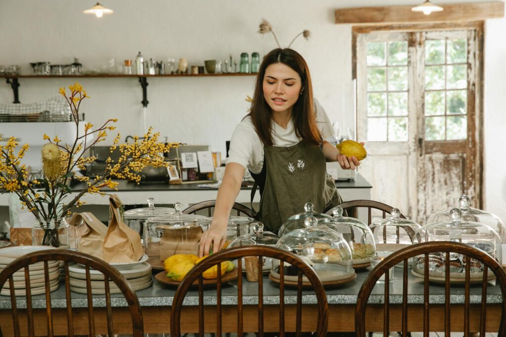 Concentrated waitress in apron standing and serving table with various delicious cakes in spacious cafe in daytime