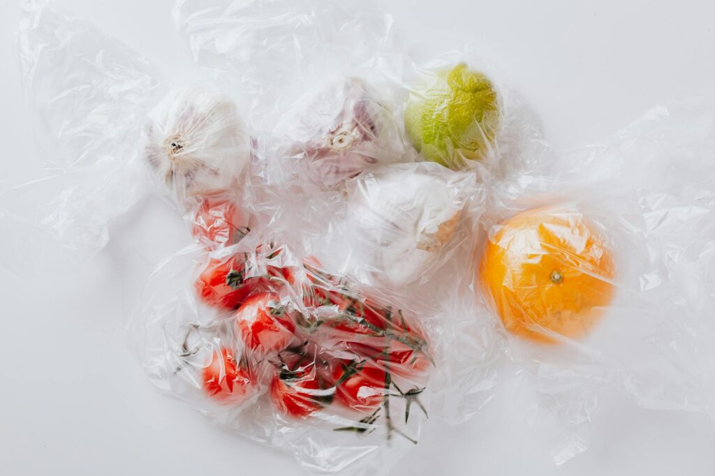 From above of bunch of ripe tomatoes put near garlic bulbs and citrus fruits covered with transparent plastic bags on white surface