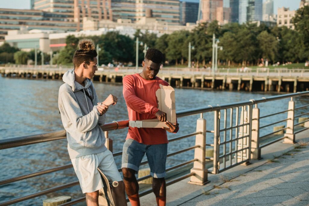 Positive diverse male friends wearing casual clothes sharing delicious pizza while standing near embankment handrail in urban city