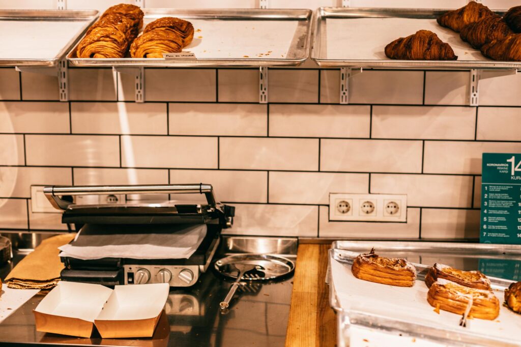 Assorted tasty puffs and croissants on metal trays above electric grill on table in cafeteria