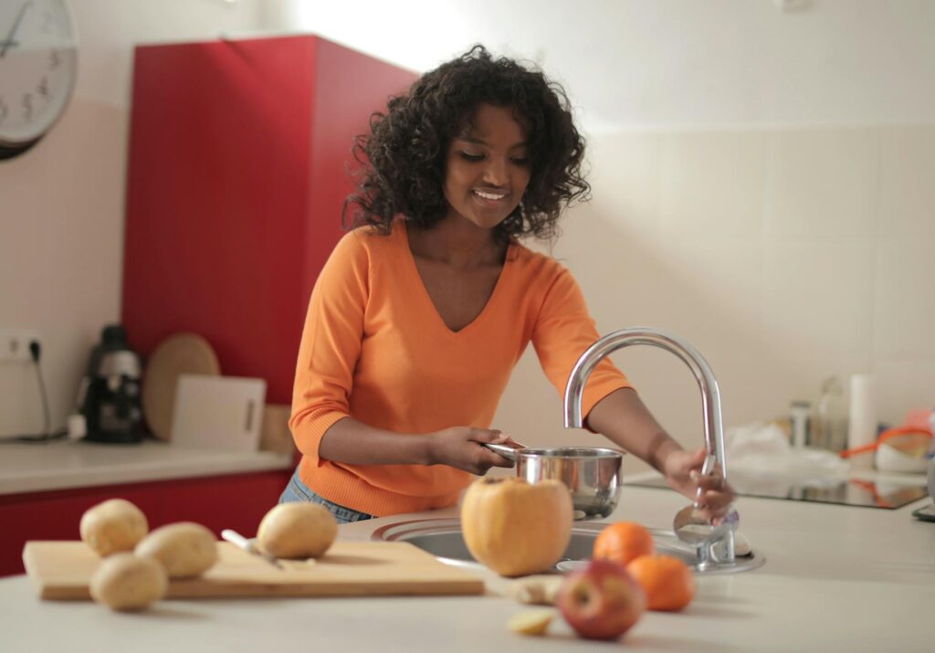 Smiling young ethnic lady in colorful casual clothes standing near sink and pouring water in pot while cooking food in spacious modern kitchen at home