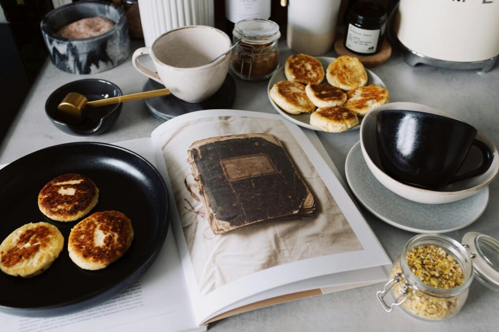 From above view of black plate with fresh cheese pancakes on opened book placed on table with various kitchenware in modern apartment